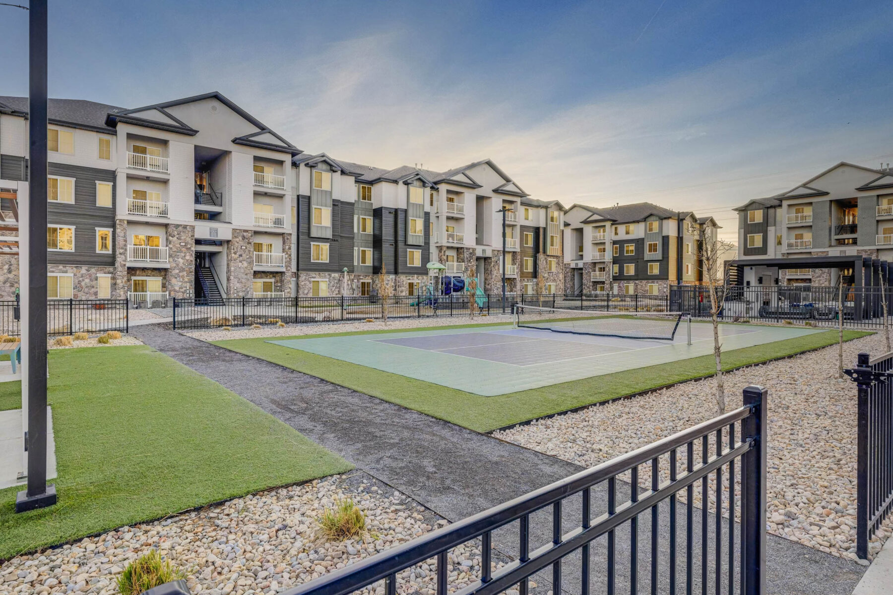 Gated outdoor recreational area with a tennis or pickleball court and a playground, surrounded by modern multi-story apartment buildings at sunset.