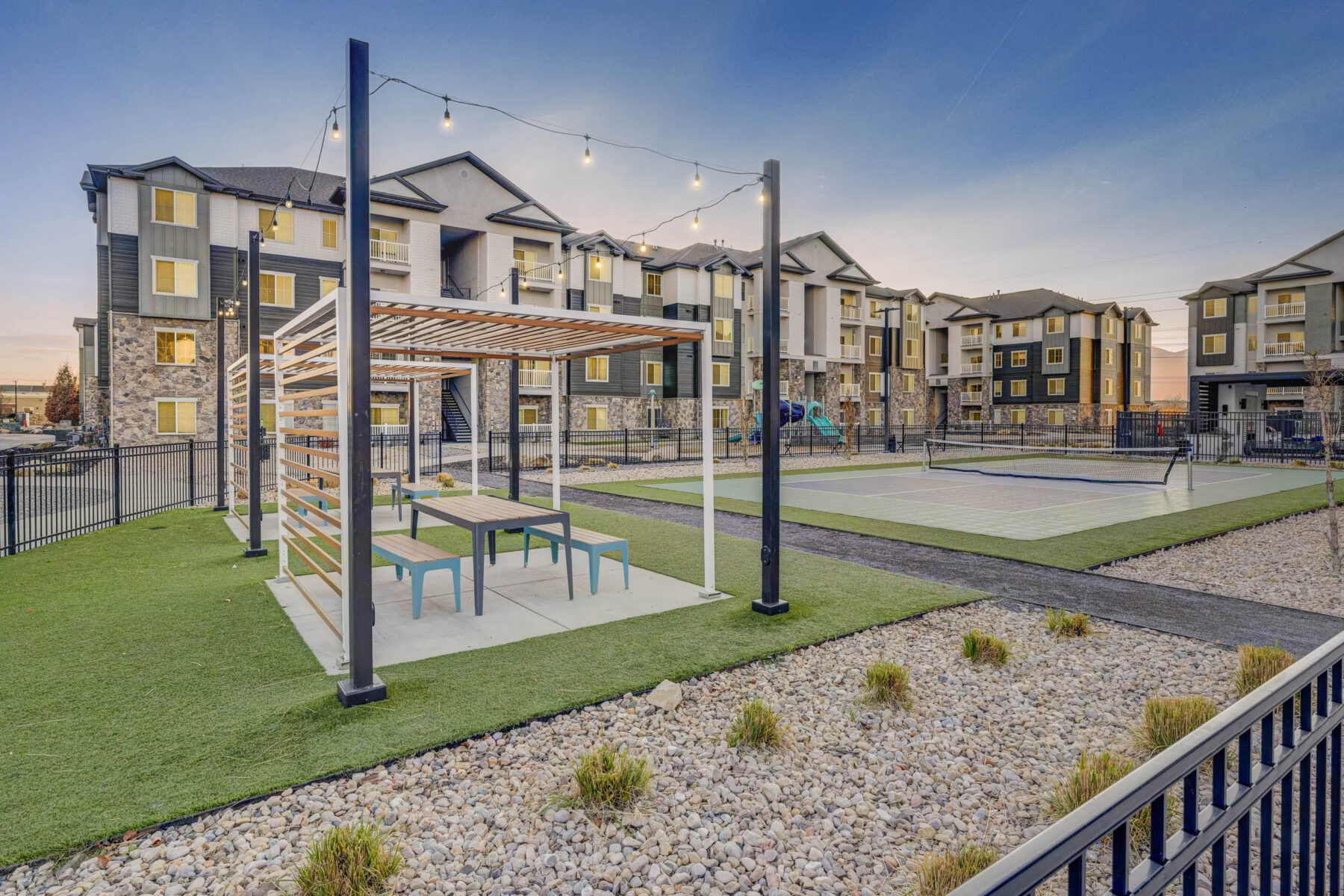 Outdoor communal area at an apartment complex featuring a pergola with picnic tables, string lights, a tennis court, artificial grass, and landscaped rocks, with multi-story apartment buildings in the background at sunset.