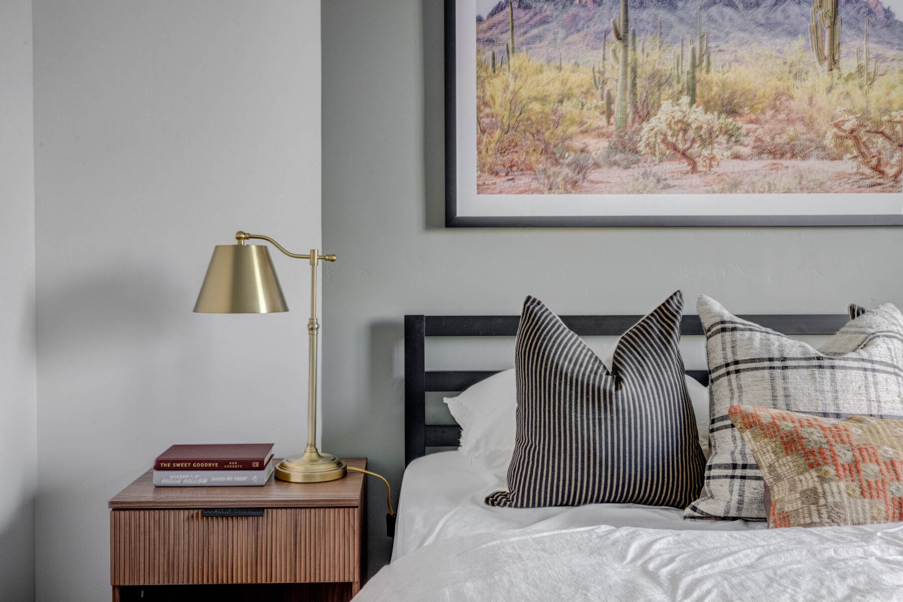 A modern bedroom features a bedside table with a brass lamp, two stacked books, and a bed with patterned pillows. Above the bed hangs a framed desert landscape with cacti.