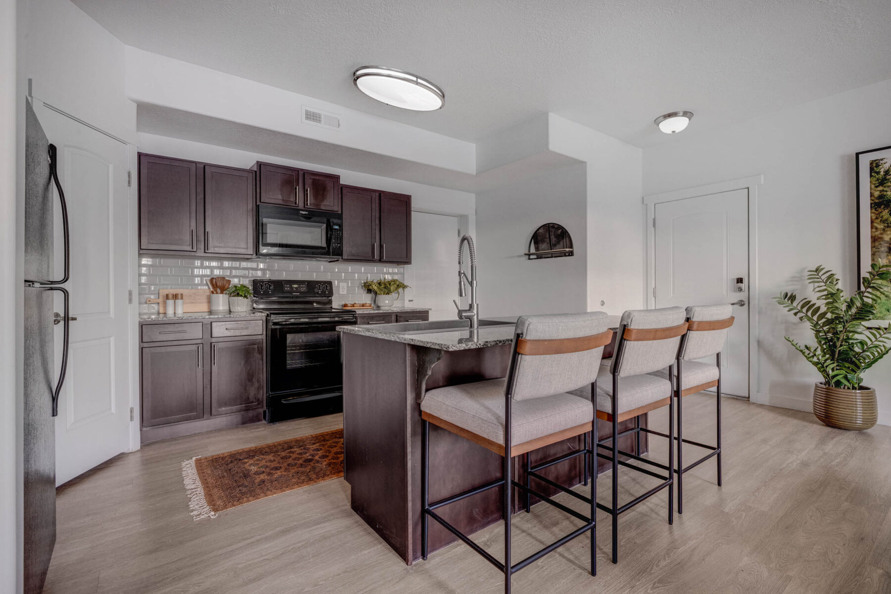 Modern kitchen with dark cabinets, black appliances, a central island with three cushioned barstools, light wood flooring, a small rug, and a potted plant near the door. White walls and minimal decor complete the space.