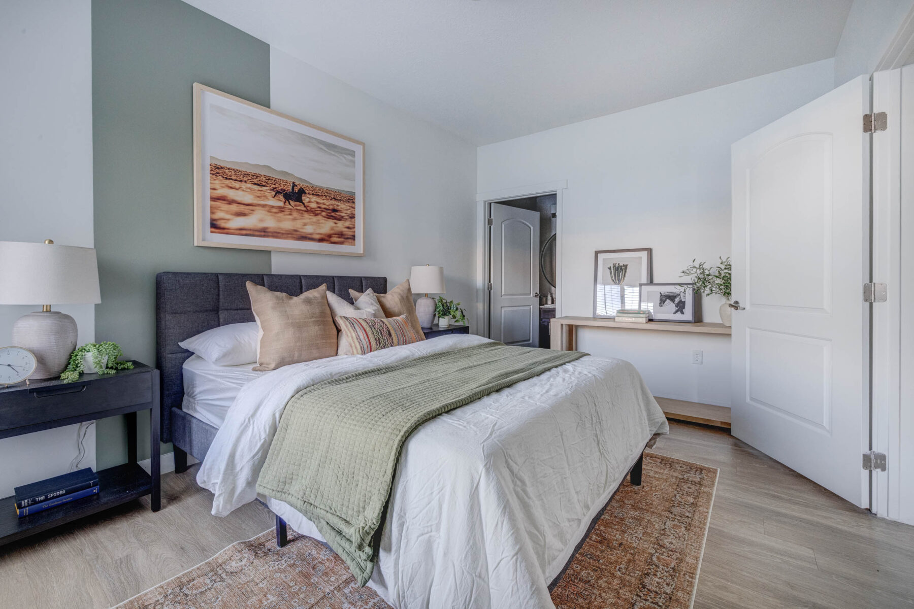 A cozy bedroom with a neatly made bed, green throw, large pillows, nightstands with lamps, a landscape photo above the headboard, and a door open to an adjoining room. Light streams in, brightening the calm space.