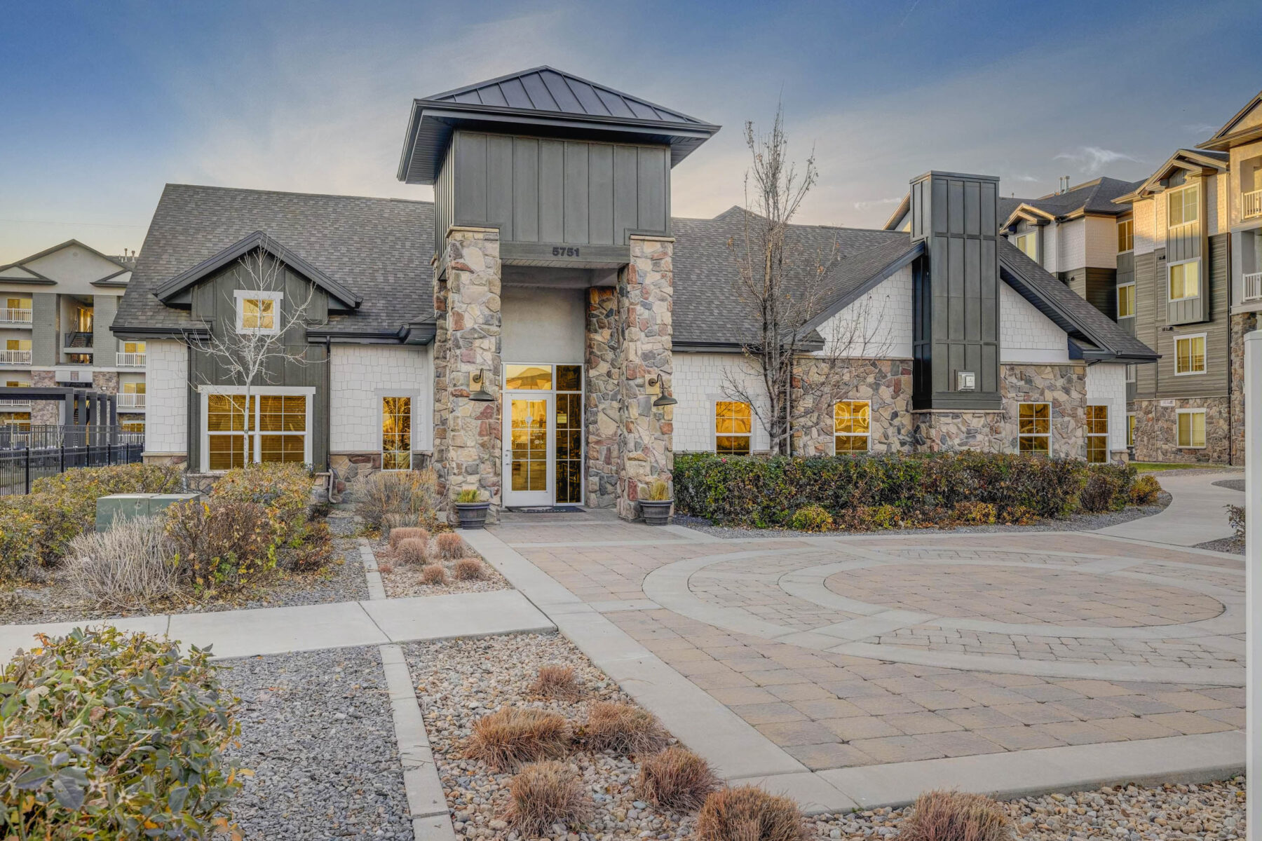 Modern apartment building with stone and siding exterior, large windows, and landscaped entrance featuring paved walkways, small shrubs, and dry landscaping under a clear sky at sunset.